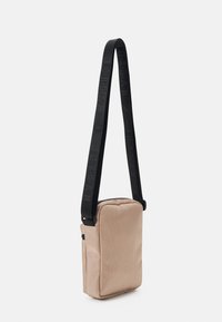 Borsa a tracolla beige con superficie testurizzata, tracolla nera regolabile con logo e chiusura superiore con zip. Forma rettangolare compatta.