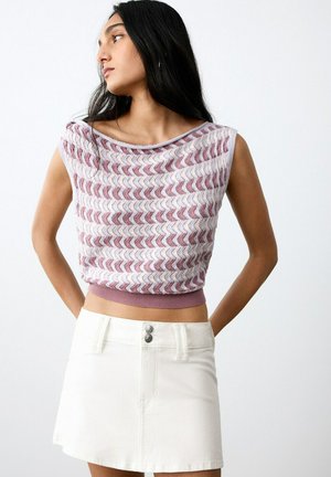 Donna con lunghi capelli scuri che indossa un top corto senza maniche con motivo e una minigonna bianca con bottoni, che guarda a sinistra.