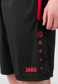 Pantalones cortos deportivos negros con acentos rojos. Presentan un logo circular y un detalle punteado en el lateral. Fabricados con un material suave y ligero.