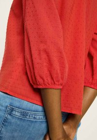 Rote Bluse mit Puffärmeln, strukturiertem Stoff mit kleinen Punkten und elastischen Bündchen. Kombiniert mit blauer Denim.