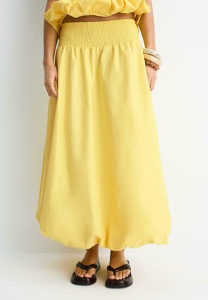 Žena, ktorá má na sebe voľnú žltú sukňu s vysokým pásom a nariaseným pásom v páse a čierne sandále s hranatou špičkou, stojaca na bielom pozadí.