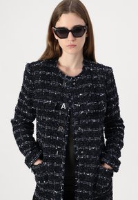 Mörkblå tweedjacka med en texturerad yta, med metallknappar, rund halsringning och två framfickor. Svarta cat-eye solglasögon bärs.
