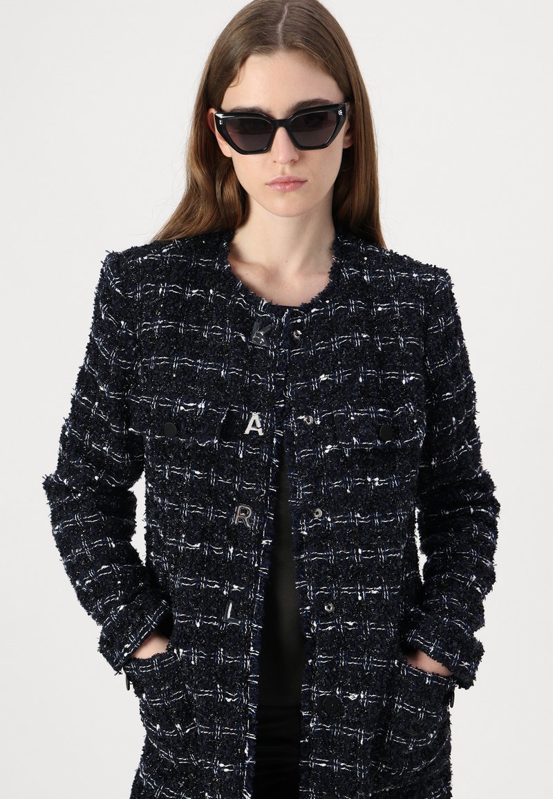 Marineblaues Tweed-Jackett mit strukturiertem Finish, ausgestattet mit metallischen Knöpfen, einem runden Ausschnitt und zwei vorderen Taschen. Es werden schwarze Katzenaugen-Sonnenbrillen getragen.