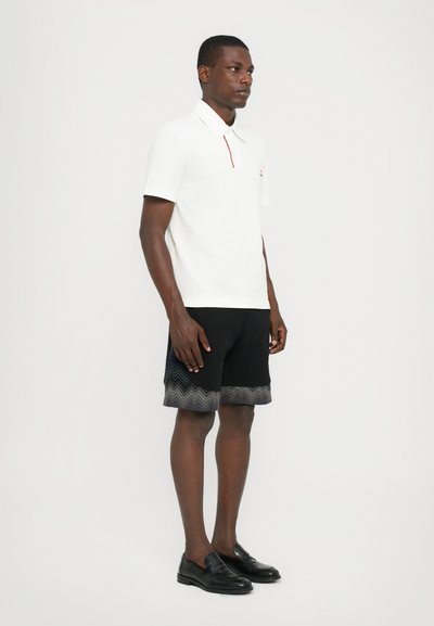 Camisa polo branca com detalhes em vermelho, combinada com calções pretos com um padrão zigzag na bainha. Sapatos loafers pretos completam o visual.