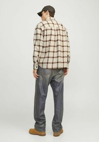 Chemise en flanelle à carreaux crème et marron, avec des poignets à boutons, associée à un jean gris ample et des bottes marron, vue de dos.
