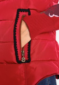 Main dans la poche zippée d'une veste matelassée rouge avec bordure noire, portant un pull en maille rouge avec un motif sur la manche.