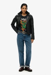 Zwarte leren motorjack, grafisch t-shirt met groene draakprint, blauwe denim jeans, gesp met studs en bordeauxkleurig schoeisel.