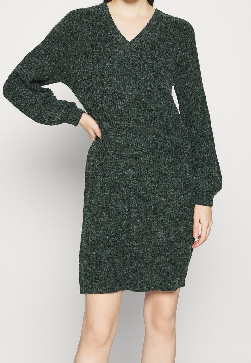 Mörkgrön stickad klänning med V-ringning och långa ärmar. Har en strukturerad mönster och en åtsittande design som slutar strax ovanför knäna.