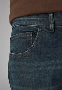 Tmavě modré džíny z denimu s přiléhavým střihem, viditelným švem a boční kapsou. Látka má strukturovaný, mírně vybledlý vzhled.