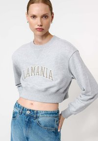 Pilkos trumpintos sportinės palaidinės su raštu „LAMANIA“, turinčios raukinėtus rankogalius ir apatinę dalį, derinamos su aukštu liemeniu džinsais.