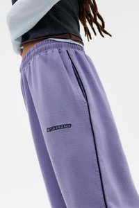Calças de fato roxas feitas de tecido macio, com uma cintura elástica, bolsos laterais e um logótipo preto bordado na coxa.