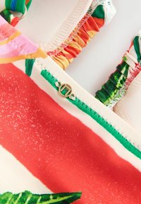 Nahaufnahme eines Badeanzugs mit bunten Stoffstreifen, grünen Nähten, einem rot-cremefarbenen Muster und einem kleinen goldenen runden Anhänger, der mit zwei Schlaufen befestigt ist.