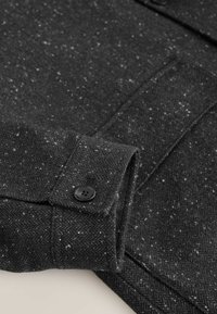 Sacou din lână cu model herringbone negru, cu finisaj texturat, având manșete cu nasturi și stropi subtili. Detaliu de cusătură vizibil.