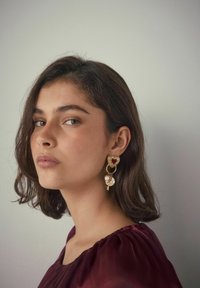 Goldene Ohrringe mit Herz- und Schlüsseldesign, mit einem roten Akzent und einem Perlendetails, die von einer runden Basis hängen. Glatte Textur.