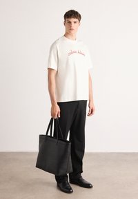 Black leather tote bag with dual handles, rectangular shape, smooth texture. Model wears a cream t-shirt with "Calvin Klein" in red script and black trousers.