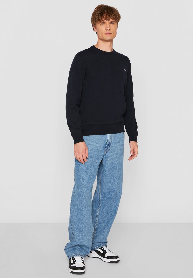 Tengerészkék kerek nyakú pulóver puha kötött anyagból, világoskék egyenes szárú farmerrel és fekete-fehér tornacipővel párosítva. Egyszerű kialakítás; minták vagy díszítések nélkül.