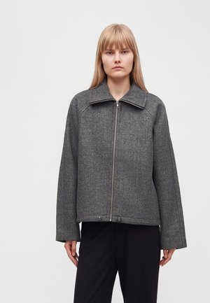 Une femme porte une veste gris charbon à fermeture éclair sur l’avant avec un large col et un pantalon noir sur un fond clair uni.