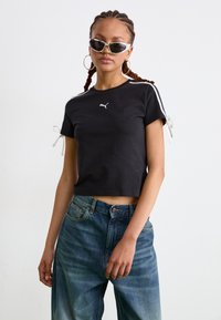  tricou scurt negru din bumbac, având dungi albe pe mâneci și accente cu fundițe, cu un mic logo alb Puma pe față.