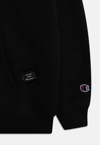 Sudadera negra con una etiqueta tejida que dice "Sé tu propio campeón" y un logotipo bordado en rojo y blanco en la manga.