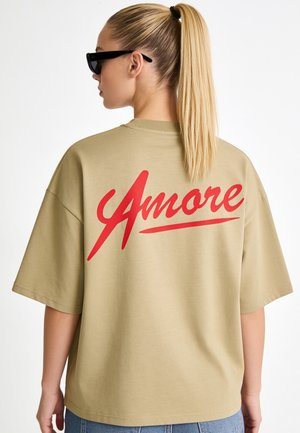 Blonde vrouw met paardenstaart draagt zwarte zonnebril en een beige oversized T-shirt met rode "Amore" tekst op de rug.