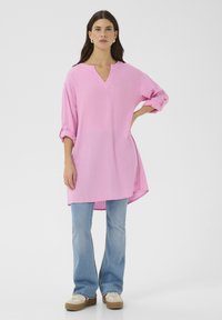 Rosa, oversize tunika med V-ringning och upprullade ärmar, i kombination med utsvängda blå jeans och beige sneakers. Enkelt mönster och mjuk textur.