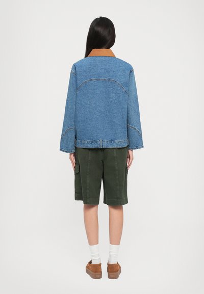 Τζάκετ από τζιν με καφέ κολάρο, χαλαρή εφαρμογή και ορατές ραφές. Συνδυασμένο με ελαιοπράσινα σορτς και καφέ σουέτ παπούτσια.