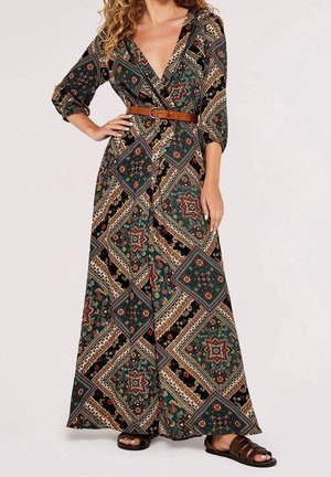 Woman wearing a patterned long wrap dress with a brown belt and brown sandals, standing with one hand on her hip.