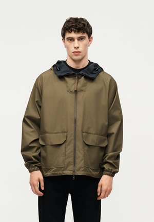 Noor mees kannab oliivrohelist lukuga jope musta kapuutsiga, elastsete mansettide ja ees olevate klapiga taskutega, seisab tühja heleda tausta ees.