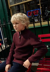 Pull en Bourgogne à col zippé avec texture côtelée, manches longues et coutures contrastantes, porté sur un pantalon bleu marine tout en étant assis sur une chaise rouge.