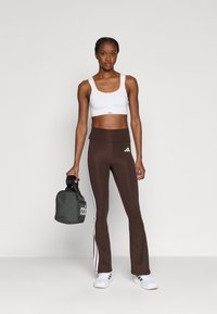 Leggings évasés marron avec des bandes blanches sur les côtés, assortis à un soutien-gorge de sport blanc. La tenue est complétée par un sac de sport gris et des baskets blanches.