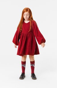 Vestido de pana rojo con mangas abullonadas largas y cintura fruncida, combinado con calcetas altas hasta la rodilla con un diseño estampado y zapatos negros con cordones.
