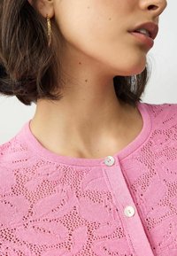Růžový pletený cardigan se květinovým krajkovým vzorem, kulatým výstřihem a dvěma knoflíky. Textura je lehká a prodyšná.