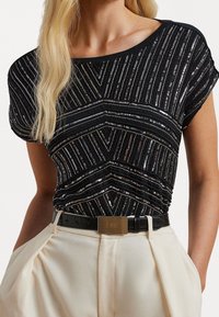 Černý top s flitry a geometrickými pruhy, z měkké látky, s širokým výstřihem a krátkými rukávy, doplněný o světle barevné kalhoty s vysokým pasem a opaskem.