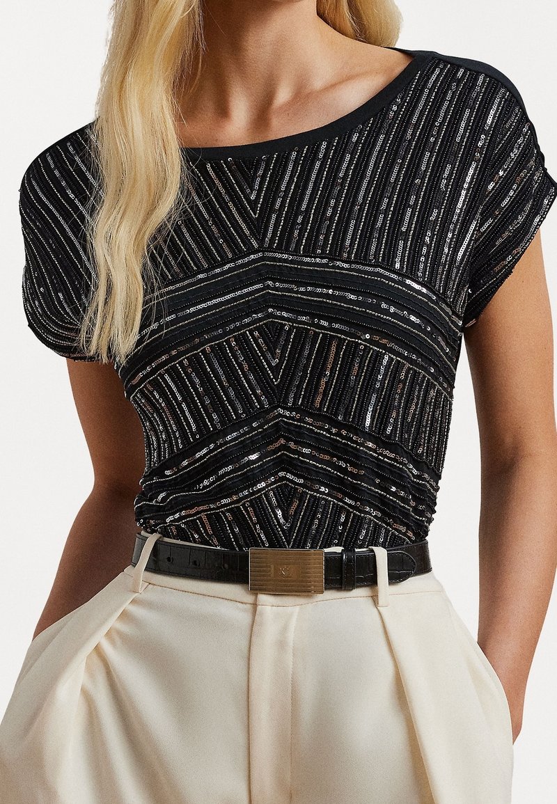 Černý top s flitry a geometrickými pruhy, z měkké látky, s širokým výstřihem a krátkými rukávy, doplněný o světle barevné kalhoty s vysokým pasem a opaskem.