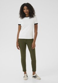 Bílý tričko s černým lemem, kombinované s olivově zelenými slim-fit kalhotami. Model má na sobě béžové tenisky s černými detaily. Hladká textura.