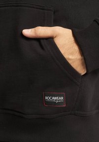 Fekete kapucnis pulóver bordás mandzsettával és aljjal, elülső zsebbel és piros fonallal varrt "ROCAWEAR" címkével egy téglalap alakú foltban.