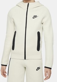Sweat à capuche Nike blanc avec fermetures éclair et logo noirs, comprenant une capuche attachée, deux poches avant et des surpiqûres contrastées. Confectionné en matière douce.