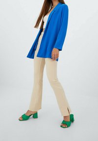 Blazer azul, top corto blanco, pantalones de campana beige y sandalias de tacón cuadrado verdes. Telas texturizadas, ajuste a medida y detalles recortados.