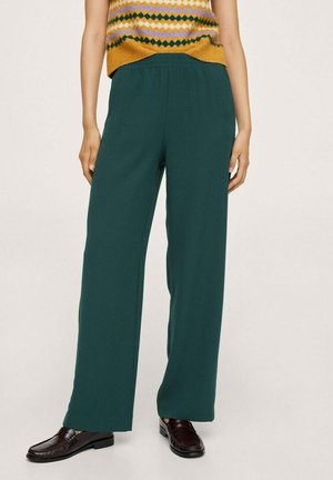 Personne portant un pantalon large vert foncé, des mocassins marron, et un pull sans manches moutarde à motifs, debout devant un fond uni.