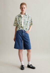 Blumenhemd mit kurzen Ärmeln in Gelb, Blau und Grün; darunter ein weißes T-Shirt. Locker sitzende blaue Jeansshorts, schwarze Schuhe und weiße Socken.