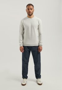 Suéter gris claro de cuello redondo con un acabado texturizado, combinado con pantalones de mezclilla azul oscuro y zapatillas blancas en una pose de pie.