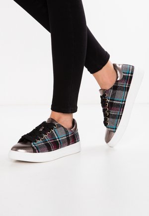 Benedenbenen in zwarte broek met geruite stofsneakers met metalen neus- en hielkapjes, zwarte veters en witte zolen op een witte vloer.