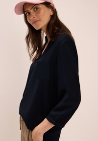 Frau mit langen braunen Haaren, die eine rosa Mütze, eine marineblaue Bluse und beige Hosen trägt, posiert mit einer Hand in ihrer Tasche vor einem beigen Hintergrund.