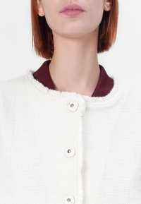 Veste en tissu blanc texturé avec un col bordeaux et cinq grands boutons blancs, présentant des bords effilochés pour le détail.