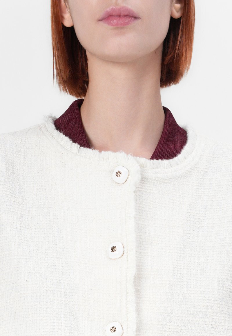 Gestikte witte stoffen jas met een bordeauxrode kraag en vijf grote witte knopen, met rafelige randen voor extra detail.