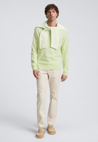 Lichtgroene gebreide trui gedragen over de schouders, crèmekleurige broek en lichtbruine loafers. Minimalistisch ontwerp met een zachte textuur.