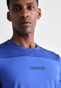 Sininen urheilupaita, jossa on verkkomateriaalia hartioilla. Edessä on musta "CALVIN KLEIN" -logo. Tasainen pinta, istuva malli.
