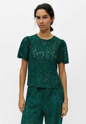 Femme portant un haut en dentelle florale vert foncé et un pantalon assorti, debout devant un fond blanc uni.