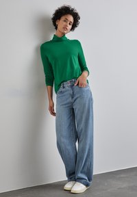 Zelený dlouhý rukáv s rolákem, spárován se světle modrými širokými džínami a bílými teniskami, na pozadí neutrální stěny.