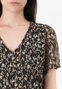 Femme portant un chemisier noir à col en V avec des manches courtes transparentes et un motif animalier abstrait beige et marron.